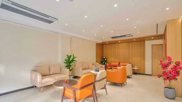 A modern hotel room with beige sofas, orange chairs, a small table, a potted plant, and a wooden panel wall.