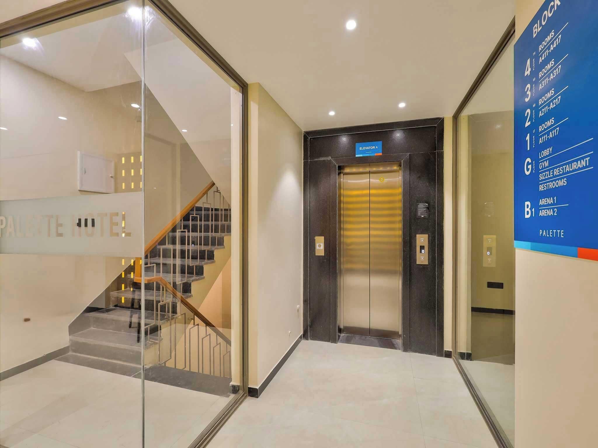 Een hotel lobby met een trap, een lift en een informatiebord waarop kamernummers en voorzieningen staan.