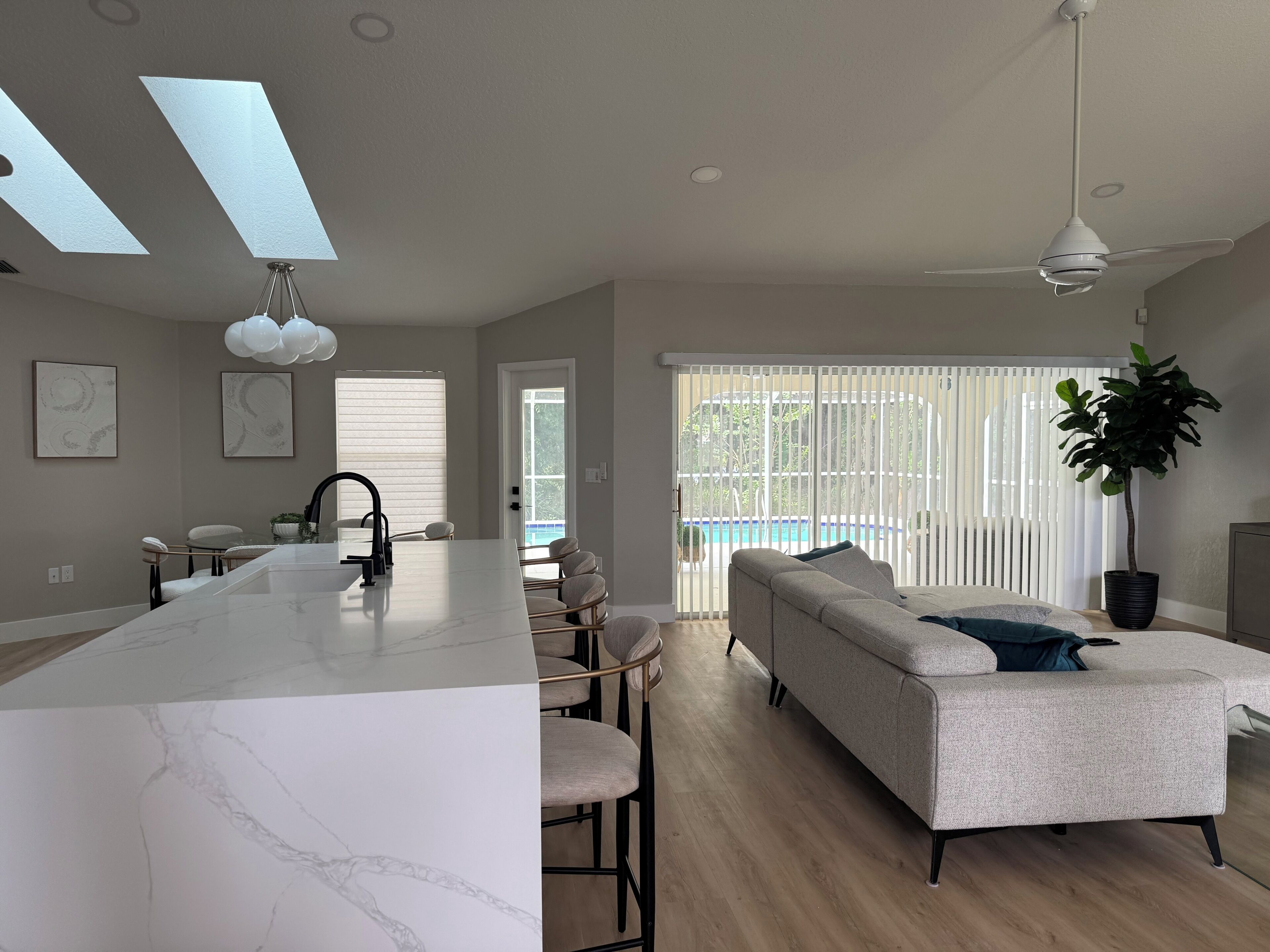 A modern kitchen with a marble island, a dining area with chairs, a sofa, and a large window with blinds.