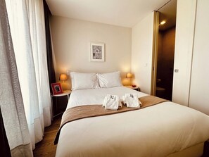Ein Hotelzimmer mit einem großen Bett, zwei Nachttischlampen, einem gerahmten Bild an der Wand und einer Tür rechts.