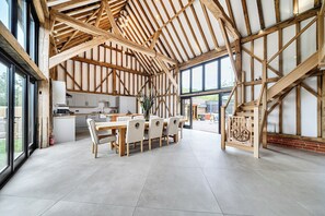 A spacious dining area with a high ceiling and exposed wooden beams, a kitchen adjacent to the dining space, and large windows providing natural light. - Barn - Sleeps 10 - Hot Tub - Parking (Reigate)