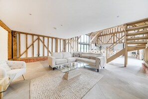 Ein geräumiges Wohnzimmer mit hoher Decke, freiliegenden Holzbalken, einem großen Fenster und einer Treppe, die in ein Obergeschoss führt.
