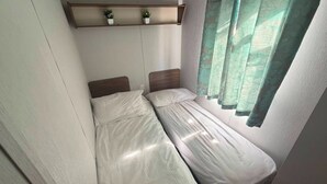 Una habitación pequeña con dos camas, un estante con plantas y una ventana con cortinas.