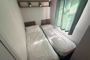 Een kleine kamer met twee bedden, een plankje met kleine planten en een raam met gordijnen.