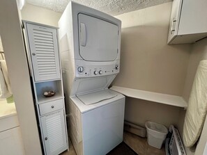 Ein kleiner Waschbereich mit einer Frontlader-Waschmaschine und -Trockner, einem kleinen Regal mit Wäschekorb und einem weißen Schrank über dem Trockner.