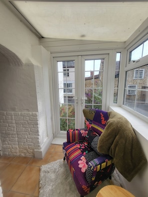 A conservatory with a chair and a rug, a stone wall, and a view of buildings outside.