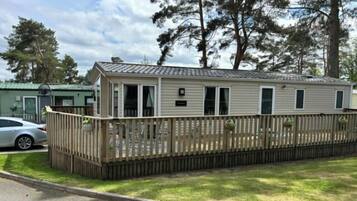 A single-story mobile home with a fenced deck, a car parked in front, and a grassy yard.
