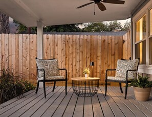 Een houten terras met twee stoelen en een tafel, een plafondventilator en een houten omheining op de achtergrond.