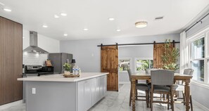 Een moderne keuken met een centraal eiland, een eethoek met een houten tafel en stoelen, en een uitzicht door een groot raam.