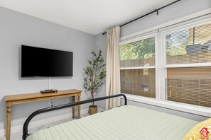 A bedroom with a bed, a television on the wall, a window with blinds, and a bedside table.