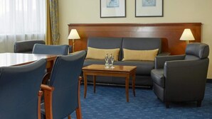 A hotel room with a sofa, two armchairs, a wooden coffee table, and a dining table. There are two lamps on either side of the sofa. The room has a large window with sheer curtains and a view of the outdoors. - 3 Person Holiday Park Home in Kassel (Kassel)
