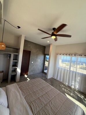 Une chambre à coucher avec un lit, un ventilateur de plafond, une fenêtre avec des rideaux, une porte et un escalier.