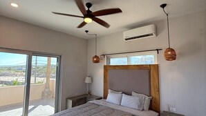 A bedroom with a bed, a ceiling fan, an air conditioning unit, a nightstand, a lamp, and a view through a large window. - Casa El Viento: Modern 3-Bedroom Desert & Sea Retreat in El Sargento (El Sargento)
