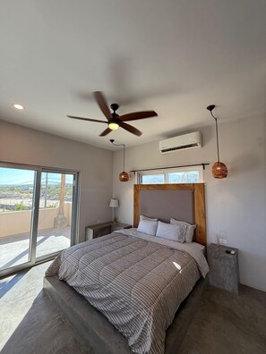 Ein Schlafzimmer mit einem Bett, einem Deckenventilator, einer Klimaanlage, einem Nachttisch, einer Lampe und Blick durch ein großes Fenster.