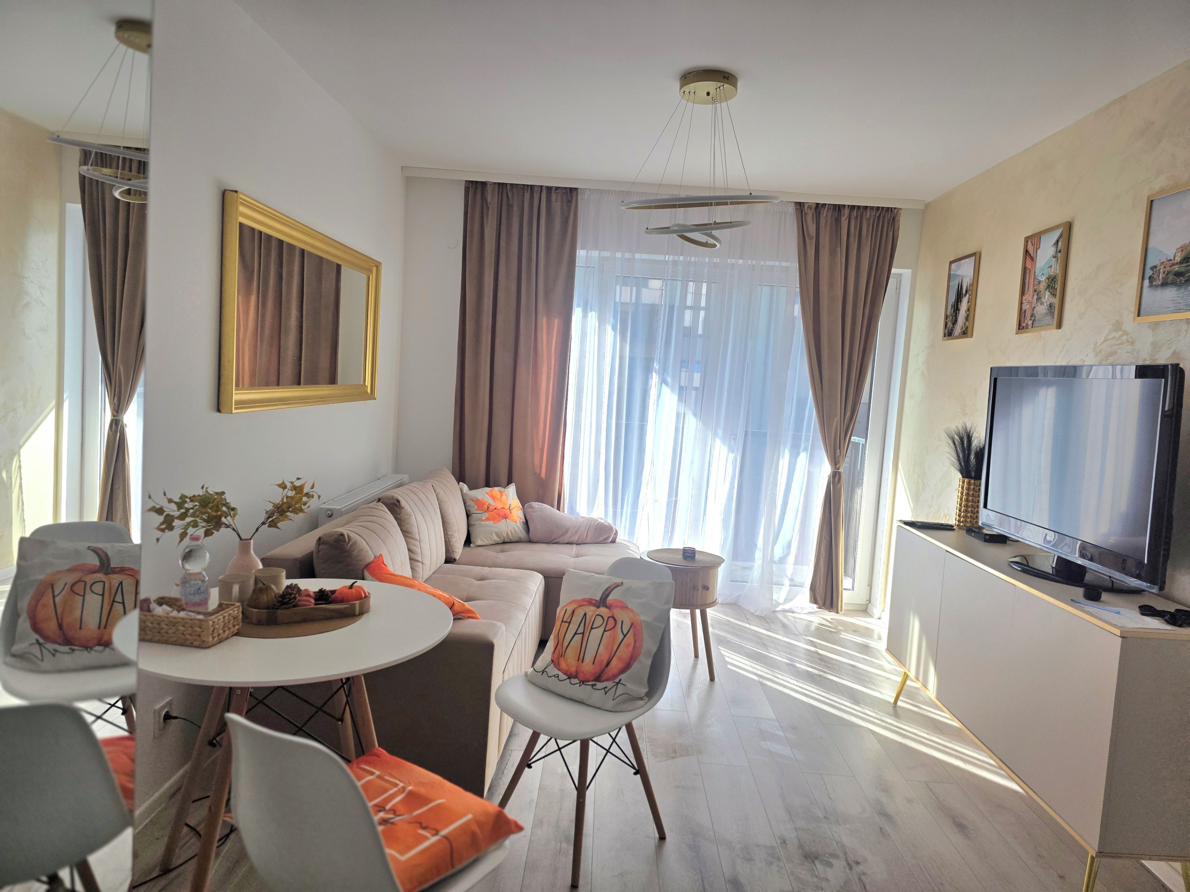 En moderne stue med sofa, spisebord, stole, fjernsyn og et stort vindue med gardiner.