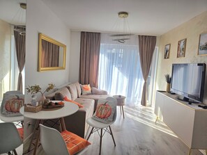 Ein modernes Wohnzimmer mit einer Couch, einem Esstisch, Stühlen, einem Fernseher und einem großen Fenster mit Vorhängen.