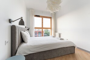 A bedroom with a bed, a bedside table, a lamp, a window, and a view of buildings outside. - Letnica Baltic Prestige Apartment (Gdańsk)