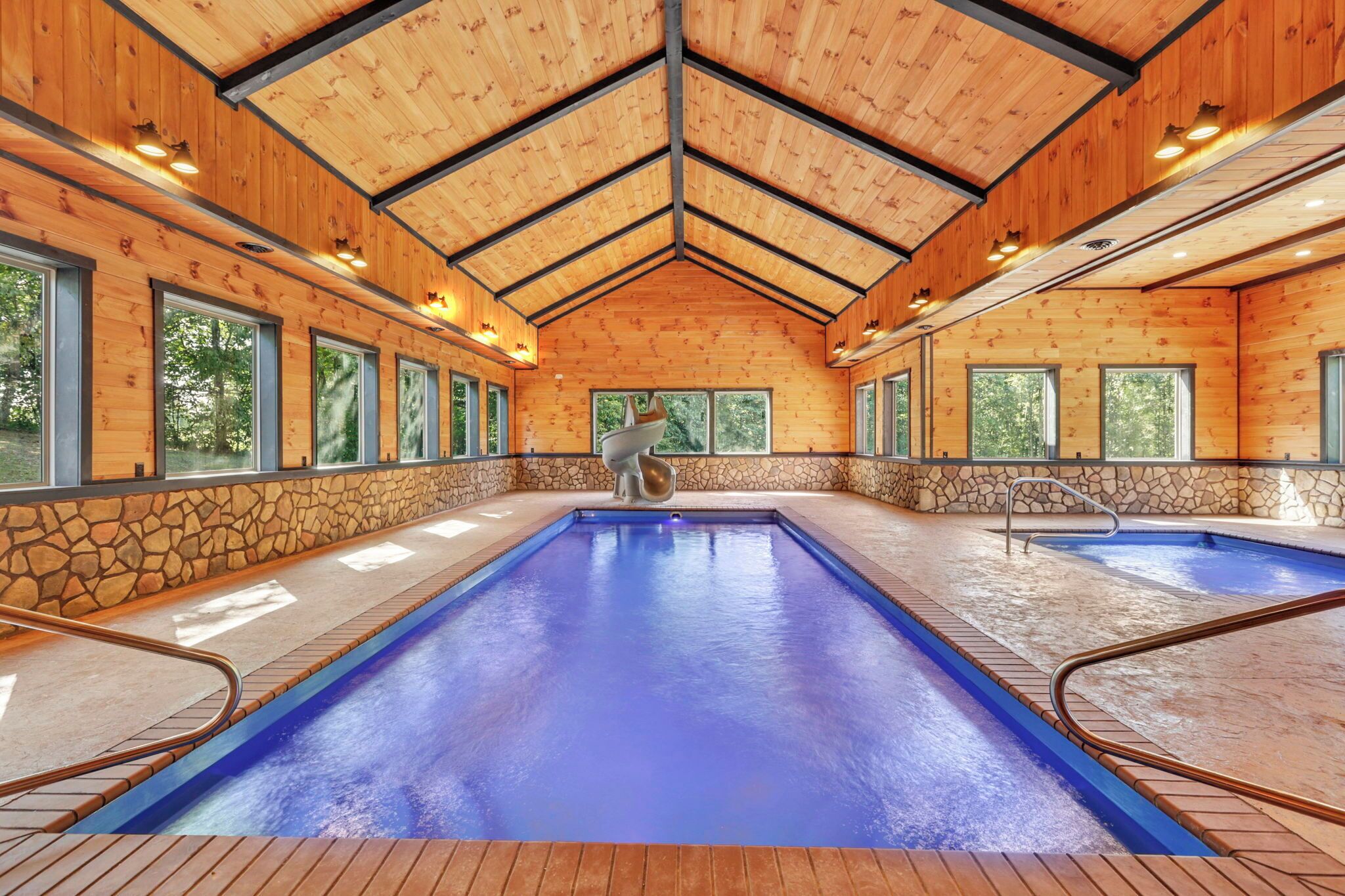 An indoor swimming pool with a stone wall and wooden ceiling, surrounded by windows.