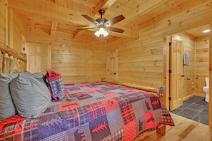 A wooden cabin bedroom with a bed, a ceiling fan, and a door leading to a bathroom. - NEW! Jenny Lynn Lodge (Logan)