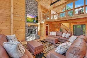 A spacious living room with a stone fireplace, leather sofas, and a large window overlooking a scenic landscape. - NEW! Jenny Lynn Lodge (Logan)