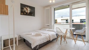 Une chambre moderne avec un lit, une petite table et une chaise. Elle possède un balcon d’où l’on peut admirer la vue sur les immeubles.