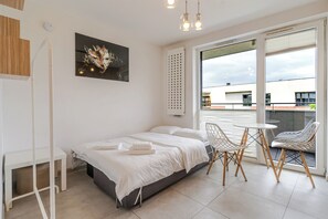 Ein modernes Schlafzimmer mit Bett, einem kleinen Tisch und einem Sessel. Ein Balkon bietet Blick auf umliegende Gebäude.