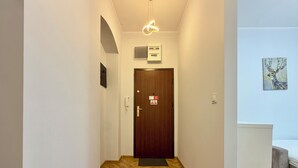 Ein Korridor mit Holzdielen im Fischgrätmuster, eine Holztür mit einem roten Schild, eine weiße Wand mit einem gerahmten Bild und ein Bett mit einer grauen Decke.