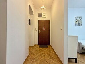 Ein Korridor mit Holzdielen im Fischgrätmuster, eine Holztür mit einem roten Schild, eine weiße Wand mit einem gerahmten Bild und ein Bett mit einer grauen Decke.