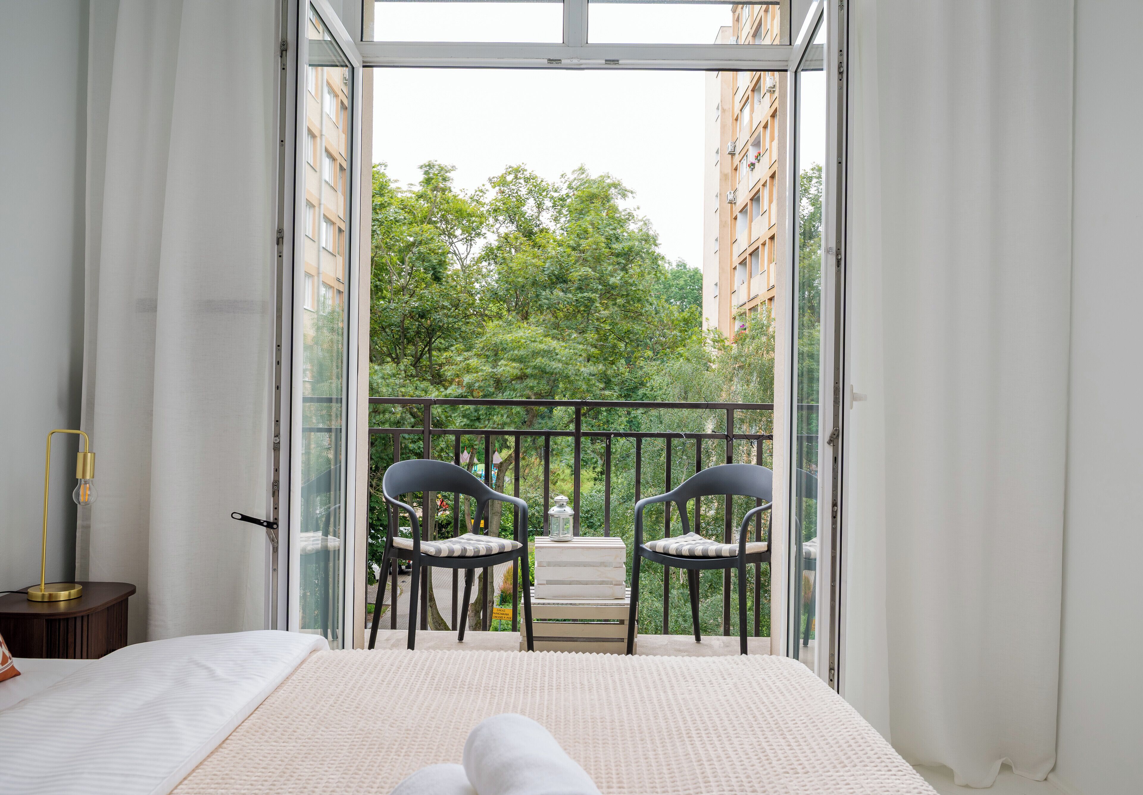 A bedroom with a bed, a bedside table with a lamp, a balcony with chairs and a table, and a view of greenery and buildings.
