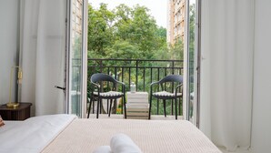A bedroom with a bed, a bedside table with a lamp, a balcony with chairs and a table, and a view of greenery and buildings. - 3 Maja Cozy Apartment (Warszawa)