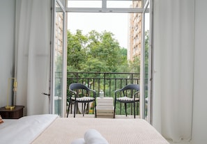 Ein Schlafzimmer mit Bett, Nachttisch mit Lampe, Balkon mit Stühlen und Tisch sowie Blick auf Grünflächen und Gebäude.