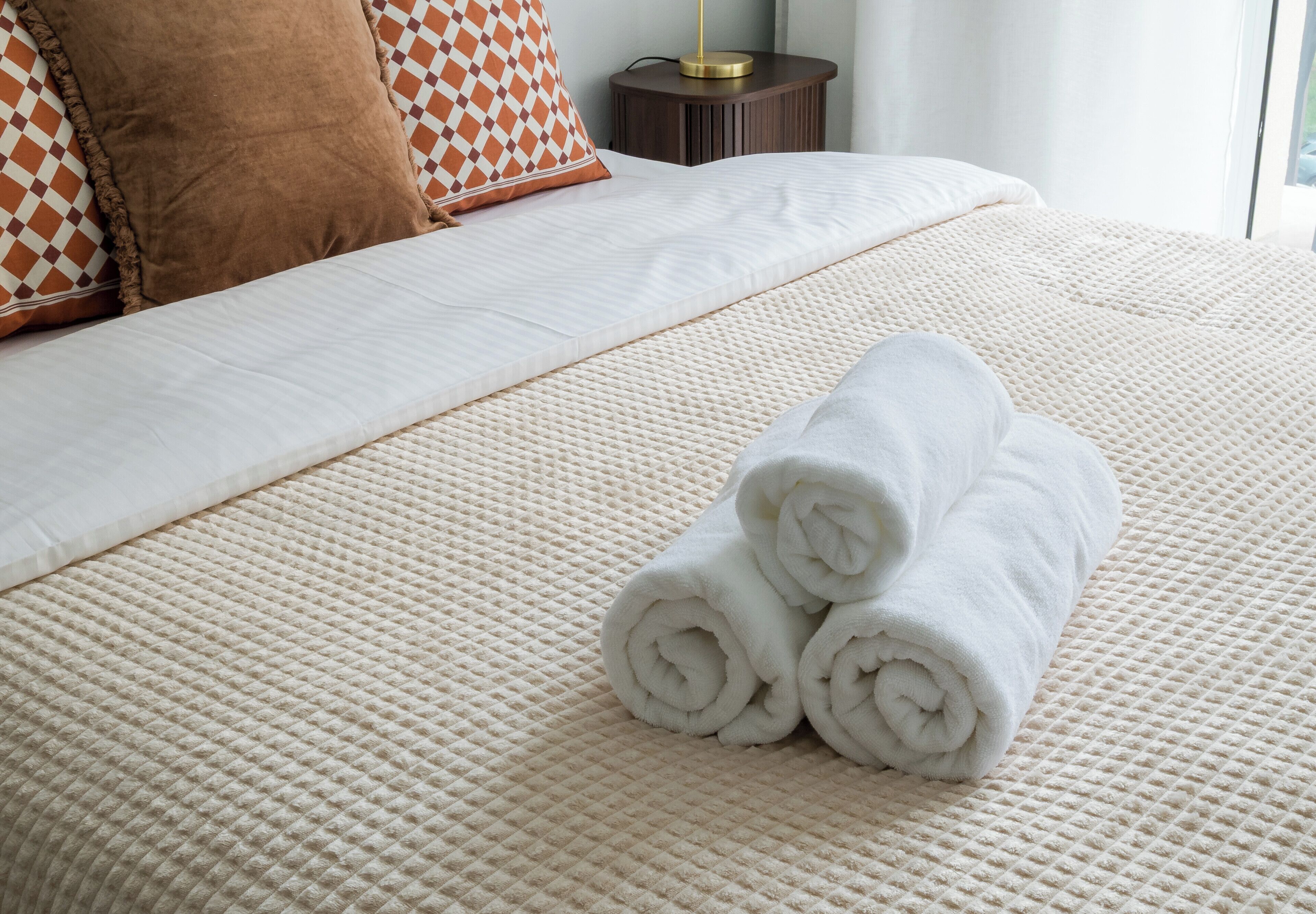 A neatly made bed with a textured white bedspread, a brown pillow, and two rolled white towels on the bed.