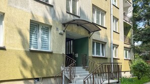 Um edifício de vários andares com escadaria que leva à entrada, grades de metal e uma pequena janela com persianas.