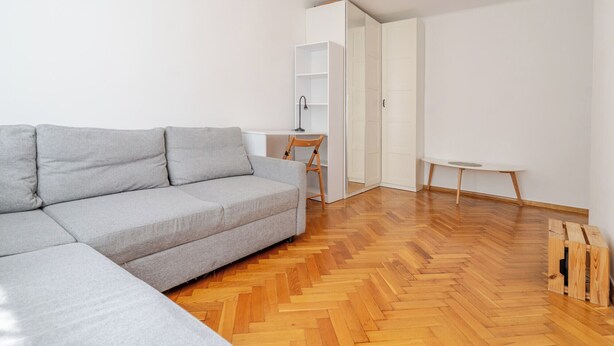 Uma sala de estar moderna com um sofá seccional cinza, piso de madeira em padrão herringbone, uma escrivaninha branca com cadeira e uma pequena mesa redonda.