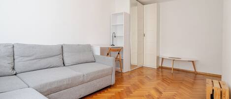 Una sala moderna con un sofá seccional gris, piso de madera en espiga, un escritorio blanco con una silla y una pequeña mesa redonda.