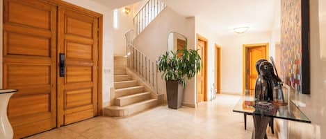 A spacious lobby with a marble floor, a staircase, wooden doors, and a glass table with decorative items.