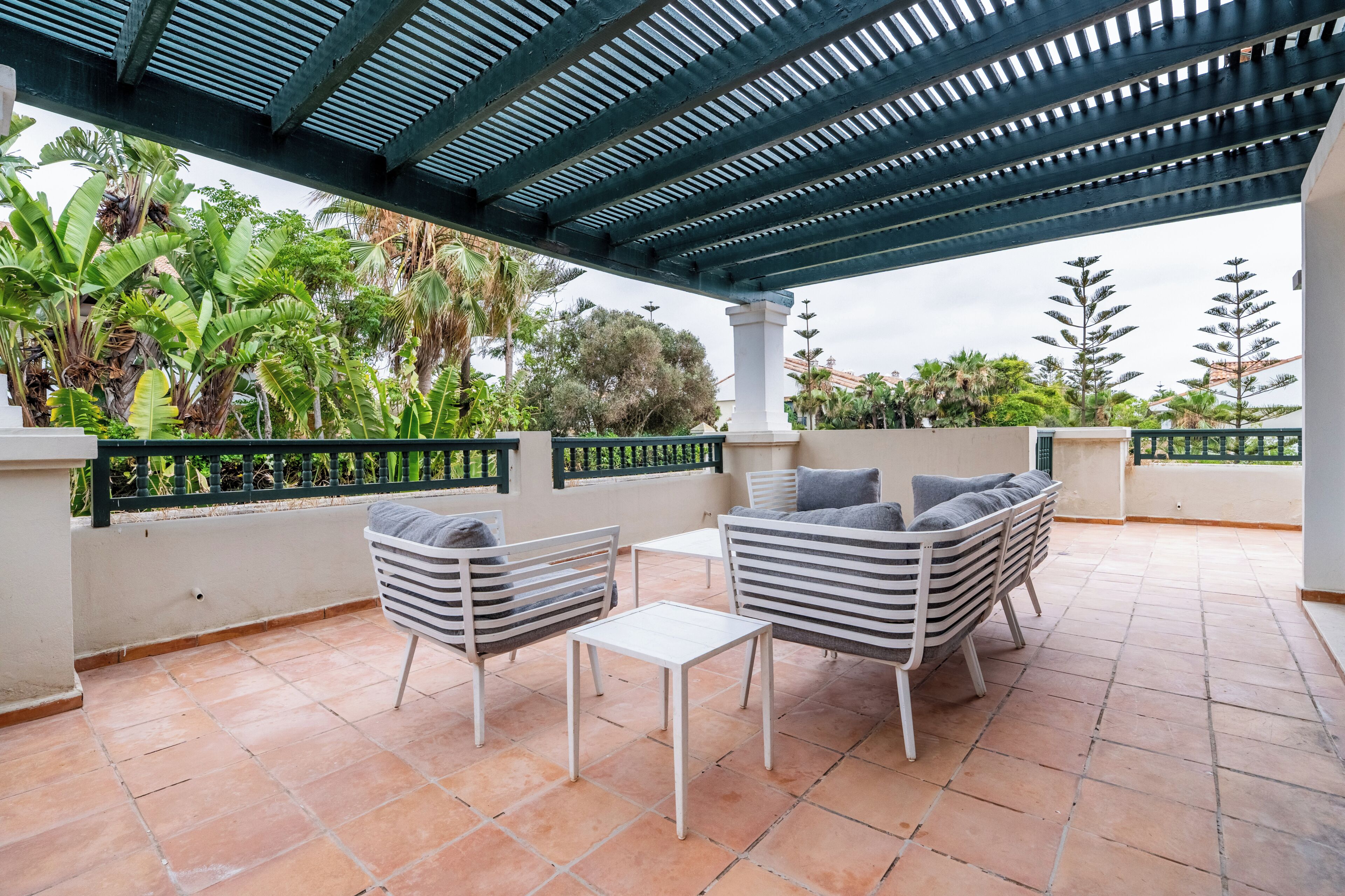 Un patio techado al aire libre con zona de estar, que incluye sillas y una mesa, rodeado de abundante vegetación y vista al cielo.