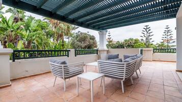 Un patio techado al aire libre con zona de estar, que incluye sillas y una mesa, rodeado de abundante vegetación y vista al cielo.