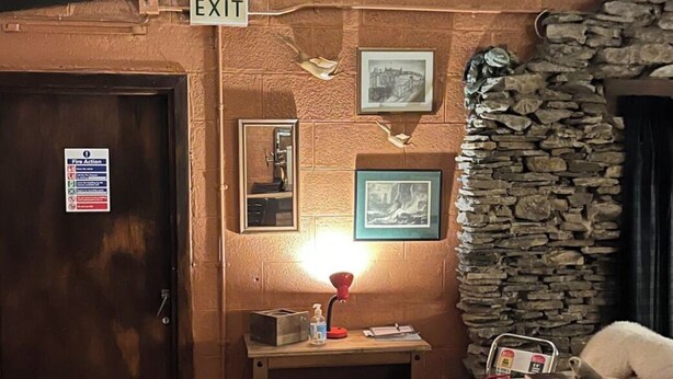 Una habitación con puerta de madera, un letrero de salida, un escritorio pequeño con lámpara, una pared de piedra y cuadros enmarcados.