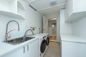 A compact kitchen with a sink, washing machine, and a small window. - Mollymere | Ocean View Coastal Retreat (Mollymook Beach)