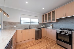 A modern kitchen with a dishwasher, stove, and wooden cabinets. - Mollymere | Ocean View Coastal Retreat (Mollymook Beach)