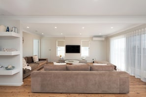 A modern living room with a large beige sectional sofa, a small round coffee table, and a flat-screen TV mounted on the wall. - Mollymere | Ocean View Coastal Retreat (Mollymook Beach)