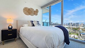 Ein modernes Schlafzimmer mit einem großen Bett, einem Nachttisch, einer Lampe und Blick auf die Stadt durch große Fenster.