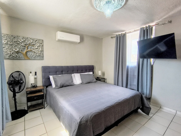 A bedroom with a bed, a flat-screen TV, a ceiling fan, a nightstand, and a wall-mounted air conditioner.