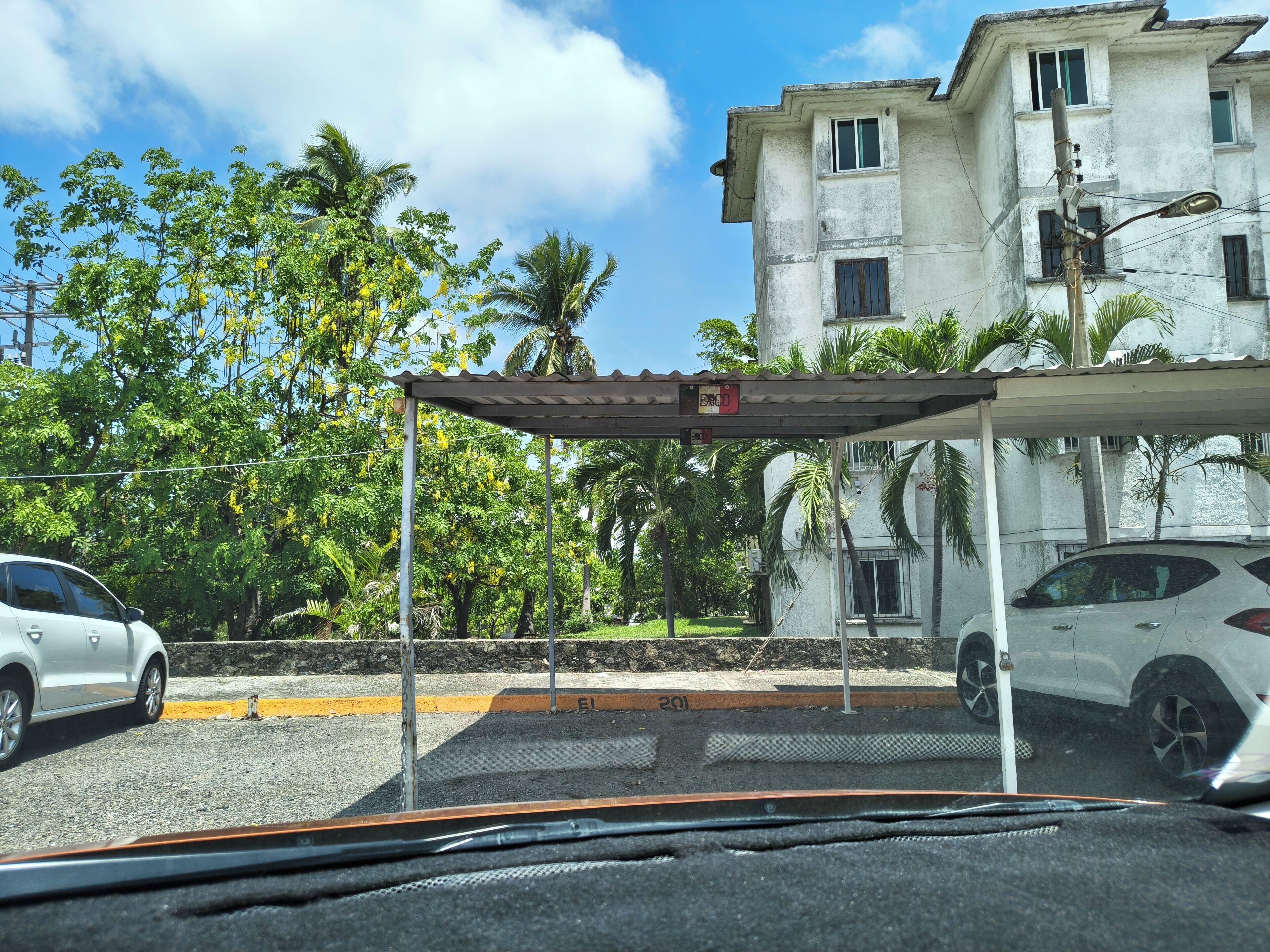 Un área de estacionamiento con un techo cubierto, un edificio con varias ventanas y palmeras al fondo.