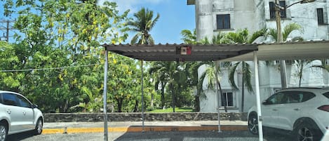 Un área de estacionamiento con un techo cubierto, un edificio con varias ventanas y palmeras al fondo.