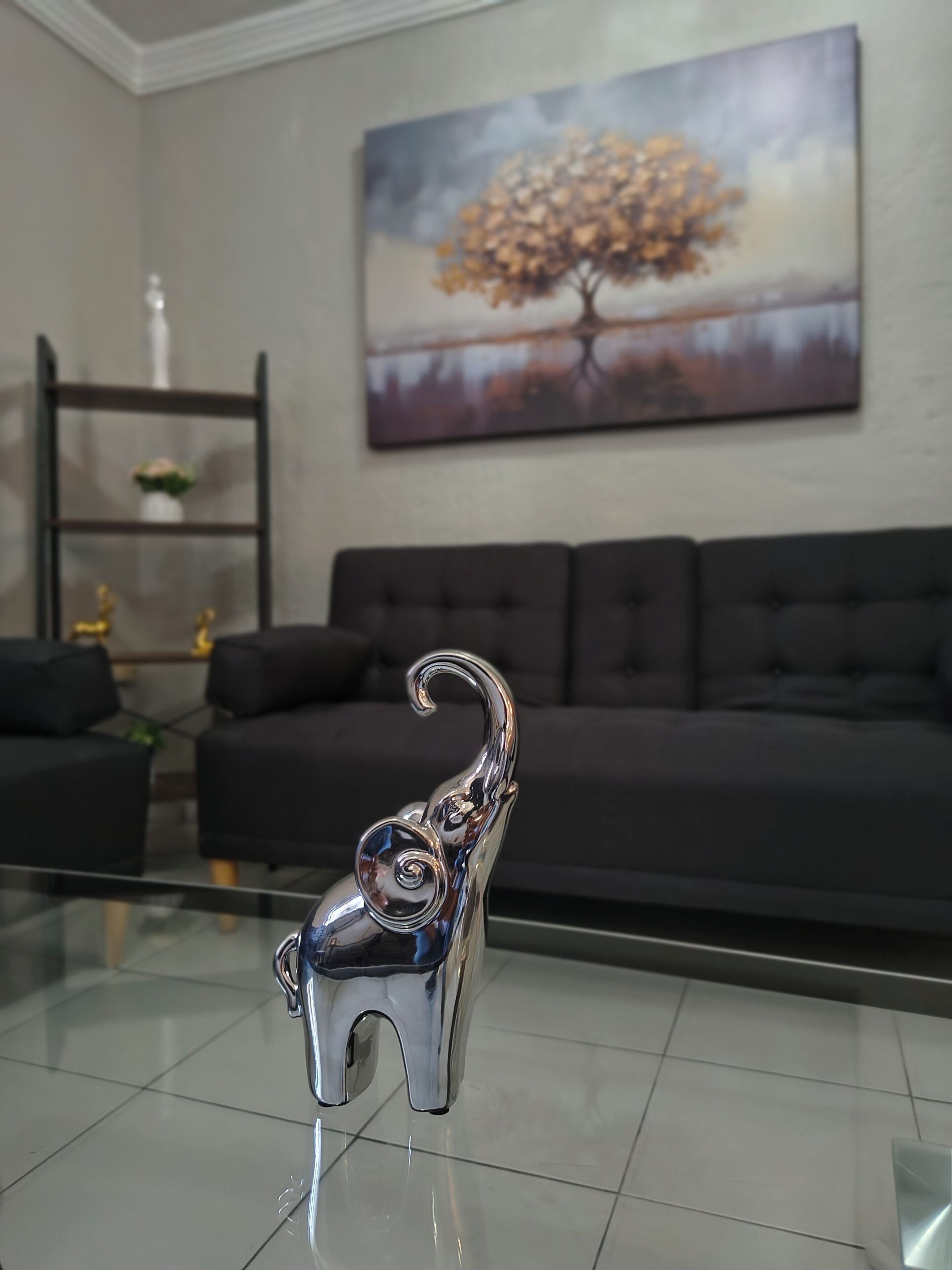 Una sala de estar con una mesa de centro de vidrio, un sofá oscuro, un cuadro de un árbol y una figurita decorativa de elefante.