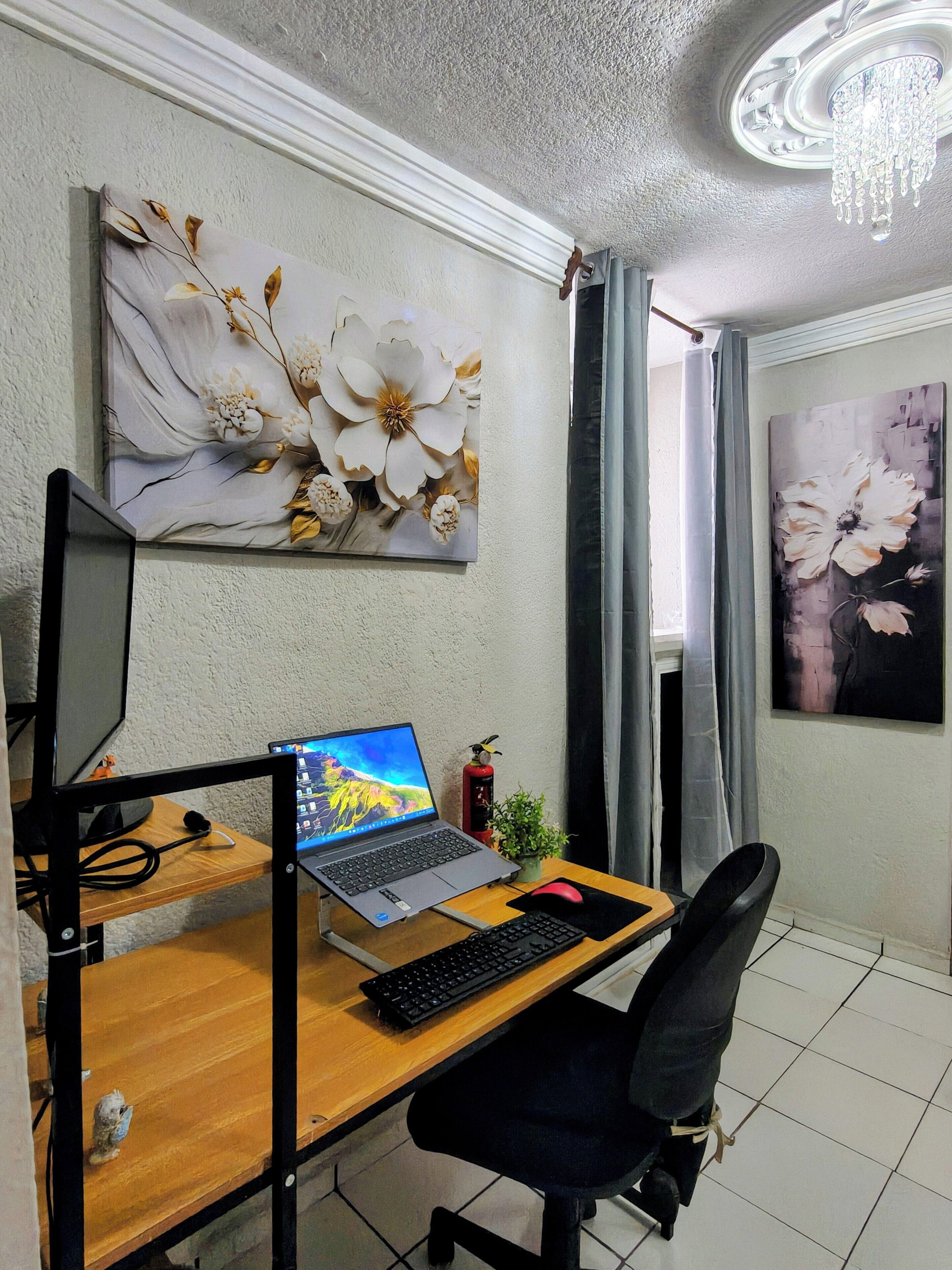 Un espace de travail aménagé à la maison, avec un ordinateur portable sur un bureau, une chaise, un écran, un clavier, une plante en pot et deux tableaux accrochés au mur.