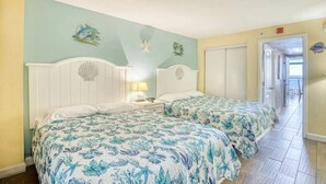Ein Zimmer mit zwei Betten, jeweils mit einem floralen Überwurf, einem weißen Kopfende mit Muscheldekor, einem Nachttisch mit Lampe und einer zur Balkon führenden Tür.