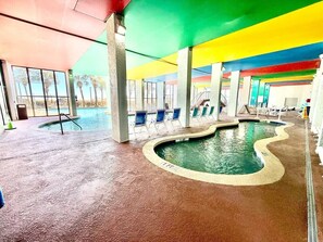An indoor swimming pool with a wavy edge, surrounded by lounge chairs and concrete pillars.